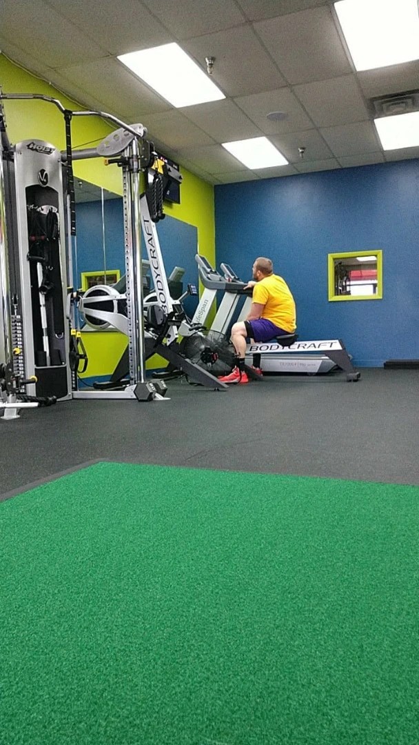 Man resting on a Bodycraft rowing machine next to green turf in a Hoffman Estates gym.