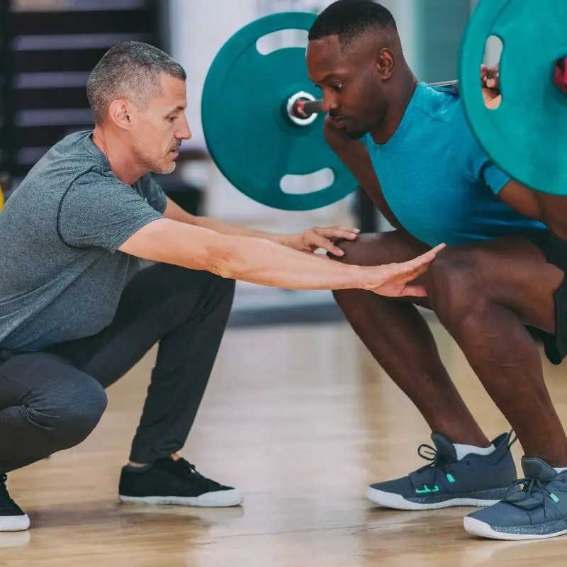 A coach provides guided strength training safety spotting for a man performing a barbell squat in a gym in Barrington, IL.