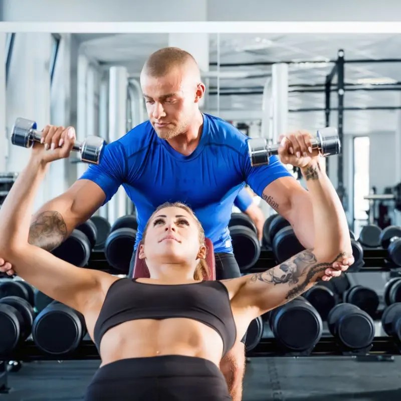 A trainer spots a woman performing a dumbbell chest press on a flat bench in Hoffman Estates, IL.